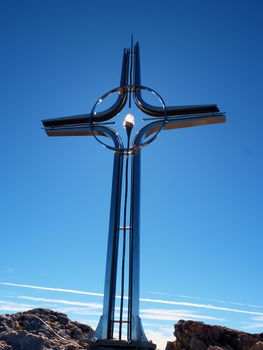 Steel cross raised  at mountain summit  in Alps. Sharp peak. Daybreak Sun in sky. Steel crucifix in memory of victims of mountains.