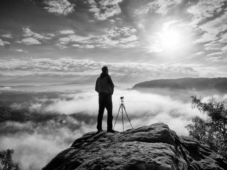 Professional photographer with tripod on cliff and thinking. Dreamy fogy landscape, blue misty sunrise in a beautiful valley below