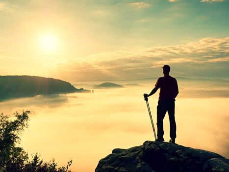 Tourist with hiking poles stand on cliff at heather bush on peak of rock empire. Man is watching over creamy fog in  valley bellow to big  Sun