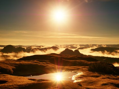 Water pool on cracked mountain summit.  Fairy daybreak in beautiful hills. Peaks of hills are in  yellow and orange fog stripped sun rays.