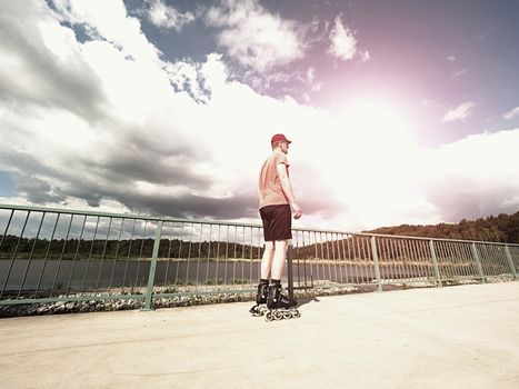 Middle age man in red t-shirt  with inline skates ride in summer park, popular outdoor roller skating. Sea bridge with blue sky in backgrround