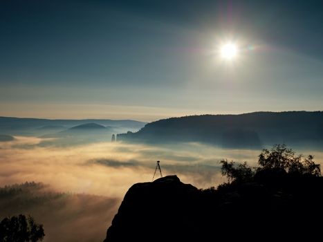 Tripod with camera on peak ready for photography. Sharp autumn rocky peaks increased from gold fog. Vivid and strong vignetting effect.