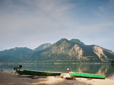 Abandoned fishing paddle boat on bank of Alps lake. Morning lake glowing by sunlight. Dramatic and picturesque scene. Mountains in water mirror.