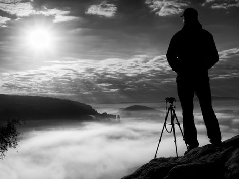 Photographer  on cliff. Nature photographer takes photos with mirror camera on peak of rock. Dreamy fogy landscape, spring orange pink misty sunrise in a beautiful valley below.