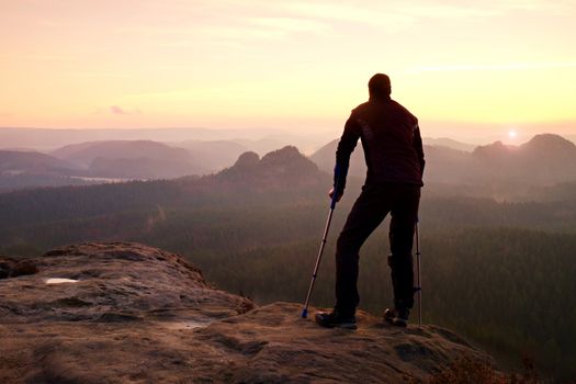 Hiker with broken leg in immobilizer. Tourist with  medicine crutch on mountain peak. Deep misty valley bellow silhouette of man with hand in air. Spring daybreak