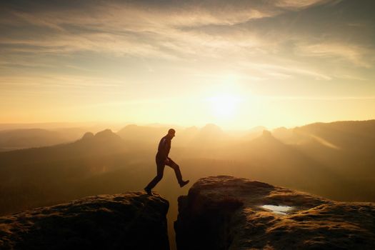 Man in black jumping between rocky empires. Dreamy daybreak in sandstone mountains, heavy orange mist in deep valley.