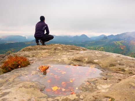 Adult tourist in black trousers, jacket and cap sit on cliff's edge looking to misty hilly valley bellow. Foggy autumn weather. Wet rock withfallen  autumn leaves
