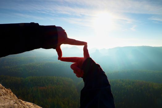 Close up  hands make frame gesture. Blue misty valley bellow rocky peak. Sunny spring daybreak in mountains.