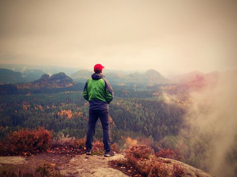 Man tourist stay on sharp rock peak. Alone hiker in red cap and green jacket  enjoy view to landscape. Vivid and strong vignetting effect.