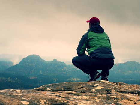 Tourist in red cap and  green black sportswear in squatting position on a rock, enjoy autumn scenery. Long valley full of heavy creamy fog after rainy night