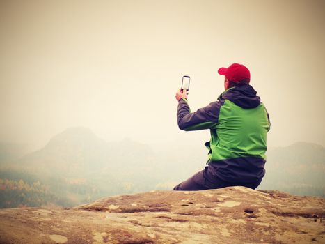 Man sitting on rock takes photos with smart phone. Hiker on peak of rock empire. Melancholy fogy landscape, spring orange pink misty sunrise in mountains.