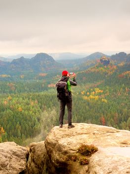 Tourist with backpack stayon cliff and  takes photos with smart phone of rainy valley. Melancholy atmosphere in foggy valley below