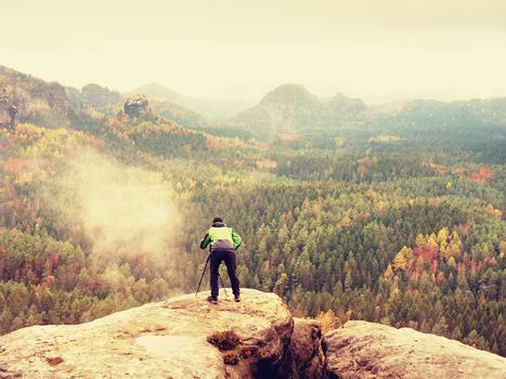 Hiker with camera on tripod takes picture from rocky summit. Alone photographer at edge photograph misty landscape, colorful forest in valley. 