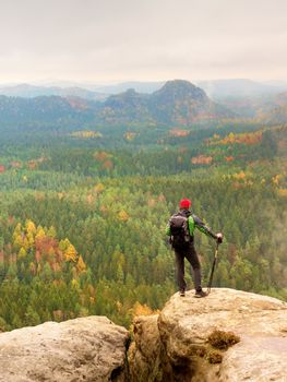 Tall hiker man with red cap,  poles in hand and with big backpack stand on rock. Misty spring daybreak in rocky mountains.