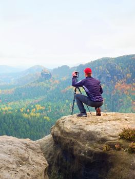 Outdoor photographer with tripod and camera on rock thinking. Creamy mist in autumnal valley bellow. Dreamy rocky mountains. Misty sunrise in a beautiful valley 