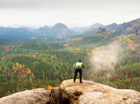 Hiker with camera on tripod takes picture from rocky summit. Alone photographer at edge photograph misty landscape, colorful forest in valley. 