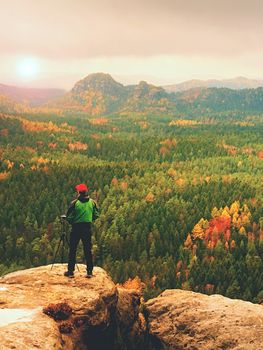 Amateur photographer prepare camera to takes impressive photos of  misty fall mountains. Tourist photographer at sharp rocky edge on high view point, colorful misty forest bellow.