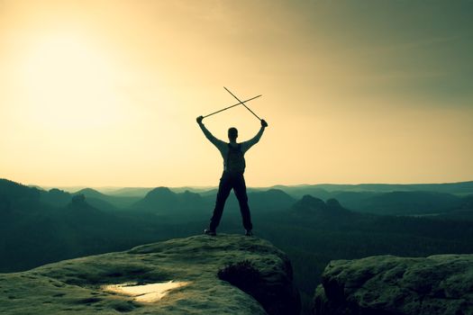 Happy hiker with x crossed poles above head.  Foggy  mountain valley bellow. Tall tourist enthusiast with poles in hands. Sunny spring daybreak in hills.