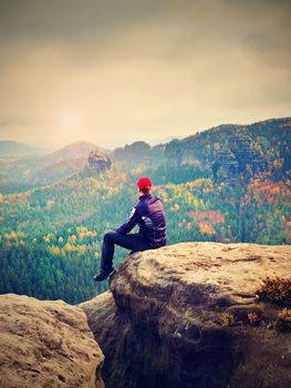 Man hiker in dark sportswear and poles stand on mountain peak rock. Exposed sandstone cliff above forest valley in national park