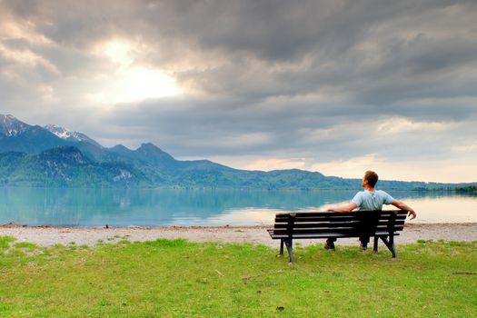 Alone  man sits on bench beside an azure mountain lake. Man relax and watch high peaks of Alps above lake mirror.