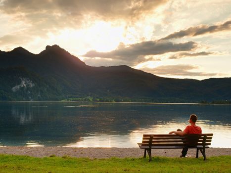 Alone man sits leg over on bench beside an azure mountain lake. Man relax and watch high peaks of Alps above lake mirror.