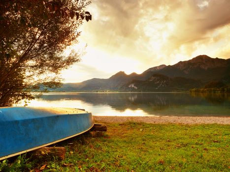Upside down fishing paddle boat on bank of Alps lake. Smooth levelof autumnal lake. Dramatic and picturesque scene. Mountains in water mirror.