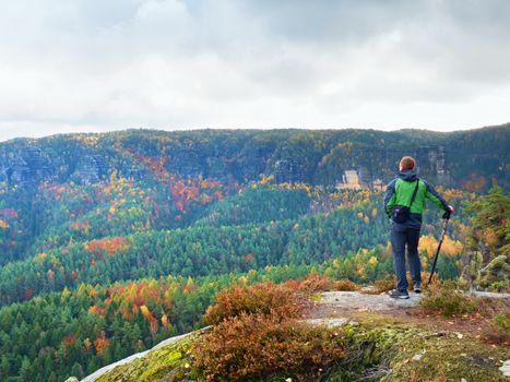 Tourist in sporty  green windcheater with poles in hands stand on rocky view point and watching into autumnal landscape. Misty spring daybreak in rocky mountains.