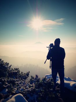 Photographer takes picture of freeze autumnal mountains, rocks covered with fresh powder snow. Stony rock peak increased from foggy valley. 