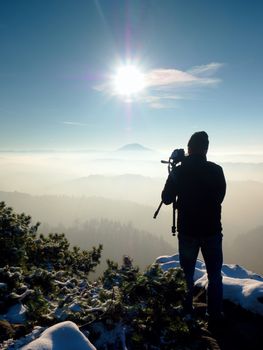 Photographer takes picture of freeze autumnal daybreak, rocks covered with fresh powder snow. Stony rock peak increased from foggy valley. 