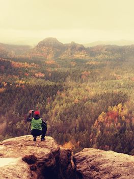 Hobby photographer takes picture of spring nature park from sharp rocks. Hiker in green jacket stay with camera on tripod on stony peak increased from foggy valley. 