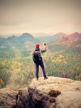 Tourist with backpack stayon cliff and  takes photos with smart phone of rainy valley. Melancholy atmosphere in foggy valley below