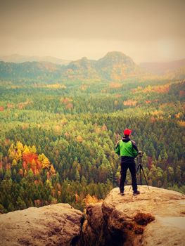 Photographer working with mirror camera and tripod on peak of rock. Dreamy fogy landscape, misty  autum day  in beautiful valley below.