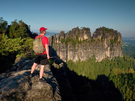 Tall tourist in red black sportswear. Sunny day in rocky mountains. Hiker with red cap, backpack and trekking poles  stand on rocky view point above green valley.