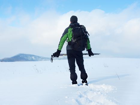 Man with snowshoes and backpack take a rest in snow. Hiker in winter jacket and trekking trousers snowshoeing in powder snow. Cloudy winter day, gentle wind brings small snow flakes 