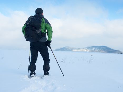 Man with snowshoes and backpack take a rest in snow. Hiker in winter jacket and trekking trousers snowshoeing in powder snow. Cloudy winter day, gentle wind brings small snow flakes 