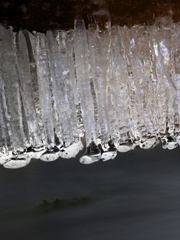 Icicles above freeze winter mountain stream. Winter season at river, shinning icicles are hanging on fallen trunk above milky water level. 