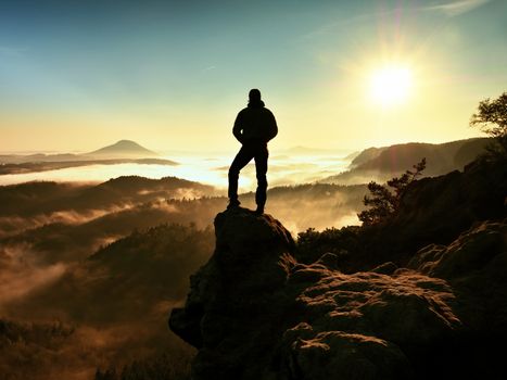 The man standing high on cliff. Hiker climbed up to rocky peak and enjoy view to valley bellow. Man  watch over misty and foggy morning valley in morning Sun. 