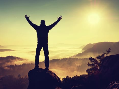 Man silhouette climbing high on cliff. Hiker climbed up to peak enjoy view. Man  watch over misty and foggy morning valley in morning Sun. 