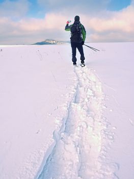 Man with snowshoes and backpack take photos by smartphone. Hiker in snowdrift,  snowshoeing in powder snow. Cloudy winter day, gentle wind brings small snow flakes 