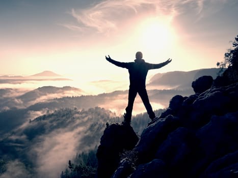 Sunny fall morning. Happy hiker with raised hands in air stand on rock above pine forest. View over misty and foggy morning valley to Sun.