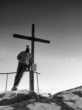 Man hiker in red blue winter clothes on sharp summit  with modest wooden cross.  Unpretentious crucifix  raised  on mountain peak in memory of victims of mountains.