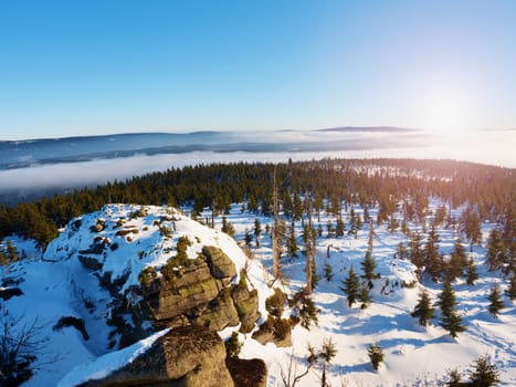 Rocky hill above inverse mist. Winter cold weather in mountains, colorful fog. Misty valley in winter mountains. Peaks of  mountains above creamy mist. 