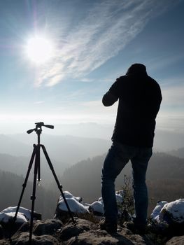 Nature photographer stay with camera ind hands  at tripod on rock and thinking. First snow. Dreamy fogy landscape, orange misty sunrise in  beautiful mountains