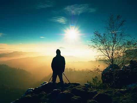 Professional photographer takes picture of freeze autumnal daybreak, rocks covered with fresh powder snow. Stony rock peak increased from foggy valley. 