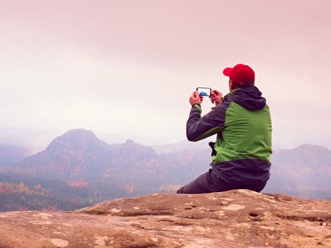 Man sitting on rock takes photos with smart phone. Hiker on peak of rock empire. Melancholy fogy landscape, spring orange pink misty sunrise in mountains.