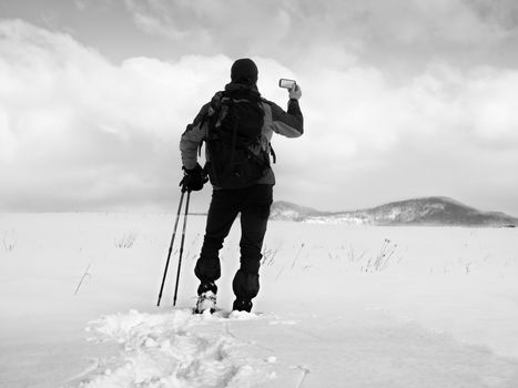 Man with snowshoes and backpack take photos by smartphone. Hiker in snowdrift,  snowshoeing in powder snow. Cloudy winter day, gentle wind brings small snow flakes 
