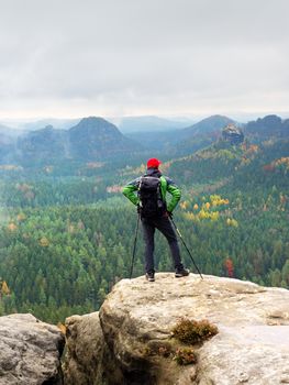 Tourist guide on the summit with poles in hand and heavy backpack. Hiker green jakcet nad red cap stand on rocky view point above misty valley. Sunny spring daybreak in rocky mountains.