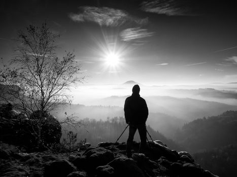 Sunny morning with first snow. Photographer preparing camera on tripod. Snowy rocks, in valley bellow colorful leaves forest. View over misty and foggy valley to Sun.