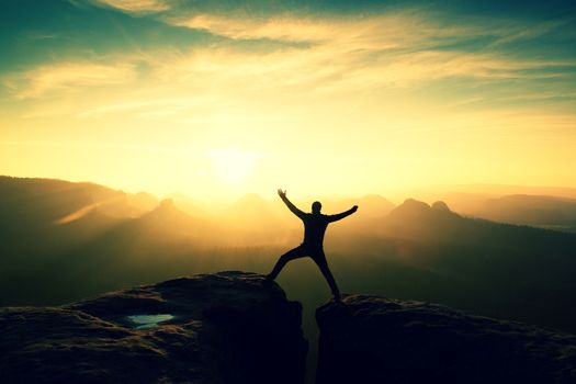 Jumping hiker in black gesture triumph between two rocky peaks. Wonderful daybreak in misty mountains.