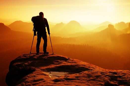 Tourist with medicine crutch above head achieved mountain peak. Hiker with broken leg in immobilizer. Deep misty valley bellow silhouette of man with hand in air. Spring daybreak